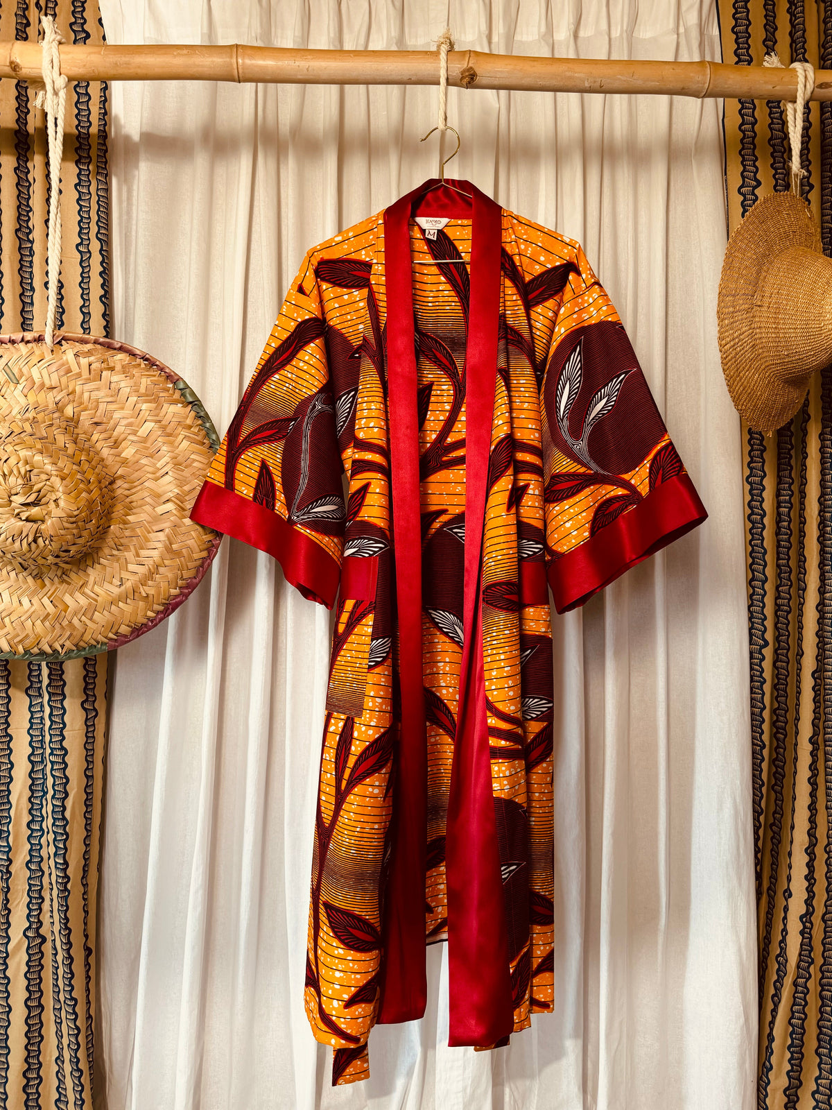 A vibrant kimono with a floral African print in shades of orange and red, with deep red satin accents, displayed on a hanging rack.